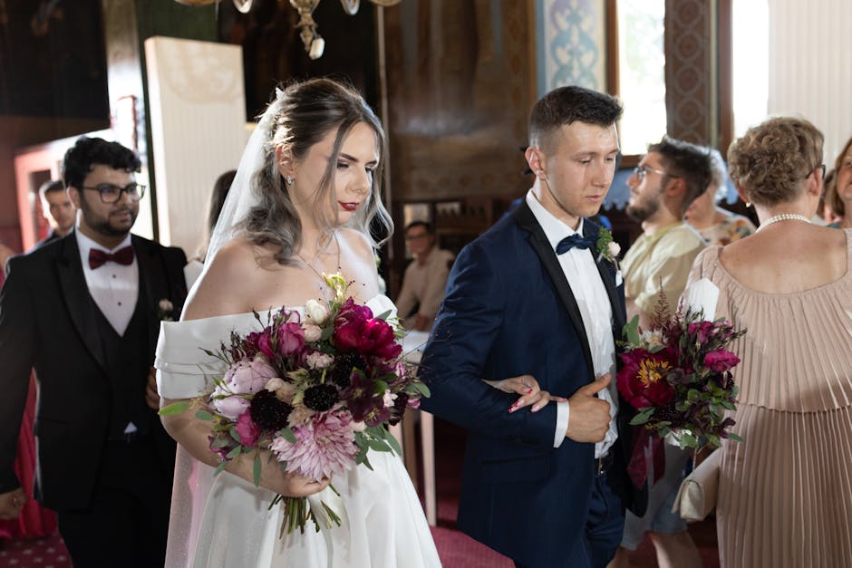 A bride and groom holding floral arrangements at a wedding ceremony inside a decorated venue. The bride's bouquet features large, fresh pink and white peonies, dark burgundy anemones, and other soft p