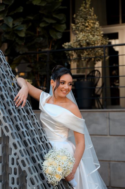 A bride in an off-the-shoulder white wedding gown, with a sheer veil over her dark hair styled in an elegant updo, is holding a bouquet of small white roses and cream-colored flowers. She is leaning a