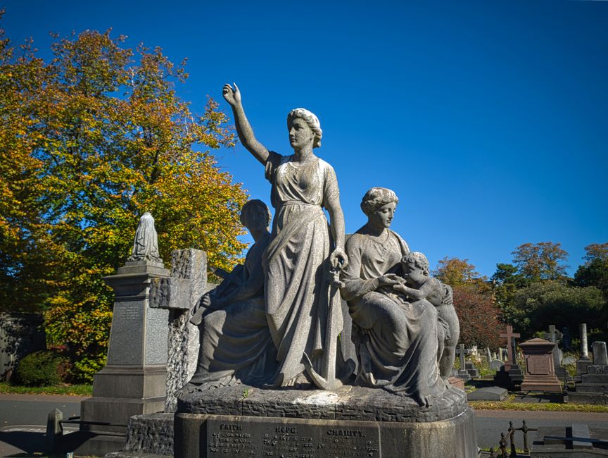 A detailed stone sculpture depicting three women in a cemetery, with one woman standing prominently raising her hand, while the other two are seated, one holding a child. The sculpture features realis