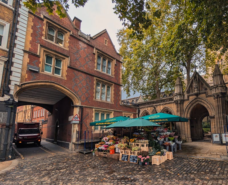 An outdoor flower market set up beneath a large green umbrella on a cobbled street corner in Earls Court SW5, featuring a variety of fresh floral arrangements and bouquets with pink, yellow, white, an