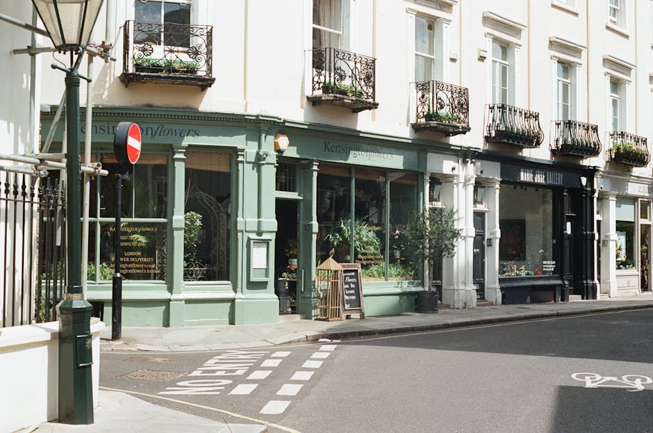 A pastel green flower shop with large storefront windows displaying various floral arrangements, located on a street corner near Earls Court station. The shop has decorative black wrought-iron balcony