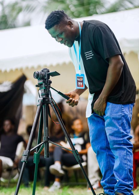The image shows a young man with dark skin and short curly hair, wearing a black T-shirt and blue jeans, standing outdoors under a white canopy tent. He is holding a smartphone in his right hand while