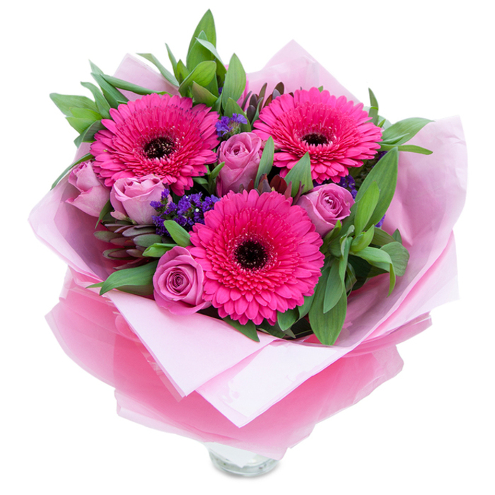 An elegantly composed pink bouquet photographed against white, anchored by three exuberant fuchsia Gerbera daisies whose concentric petals radiate out from deep, near-black centres. Interspersed among these focal blooms are clusters of lavender-tinged roses in a dusty-rose hue, their petals tightly rolled in the fresh, early stages of opening, offering a restrained, romantic counterpoint to the gerberas' boldness. Small sprays of purple statice peek between blooms, their papery texture and cool tone introducing playful contrast and dimension. A generous backdrop of glossy green leaves-likely salal or eucalyptus-creates a garden-fresh fullness and highlights the bouquet's vibrant palette. Hand-wrapped with care, the arrangement sits in an inner layer of matte pale-pink tissue and an outer sheet of sheer, slightly crinkled fuchsia-pink cellophane that catches light with a soft shimmer; the wrapper is folded with intentional creases, suggesting thoughtful presentation. The composition shows the rim of a clear glass vase at the base, inviting immediate display on a mantel or dining table. In soft morning light the petals glow; the imagined scent is a light, floral rose note with a faint green crispness. As a local florist might place this in a shop window near Earl's Court Road or tuck it into a delivery heading toward Earls Court Gardens, it reads as a versatile gift for birthdays, Mother's Day, anniversaries, or to offer encouragement-an arrangement that feels like a warm, floral embrace.